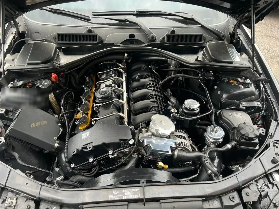 Engine bay of a BMW E90 3-series with an aftermarket intake, a gold coil-pack bracket and polished charge piping