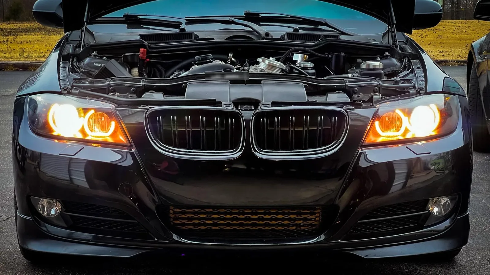 Black BMW E90 3-series parked outdoors with the hood open and its halo headlights on